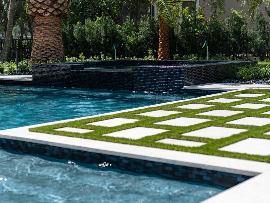 Raised spa wall clad in dark glass mosaic tile beside a pool with a paver-and-turf deck