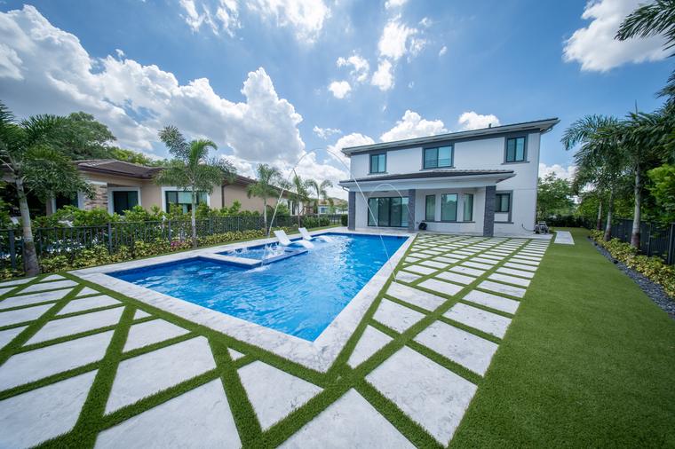 Rectangular pool with a raised spa, travertine and turf deck behind a white two-storey home